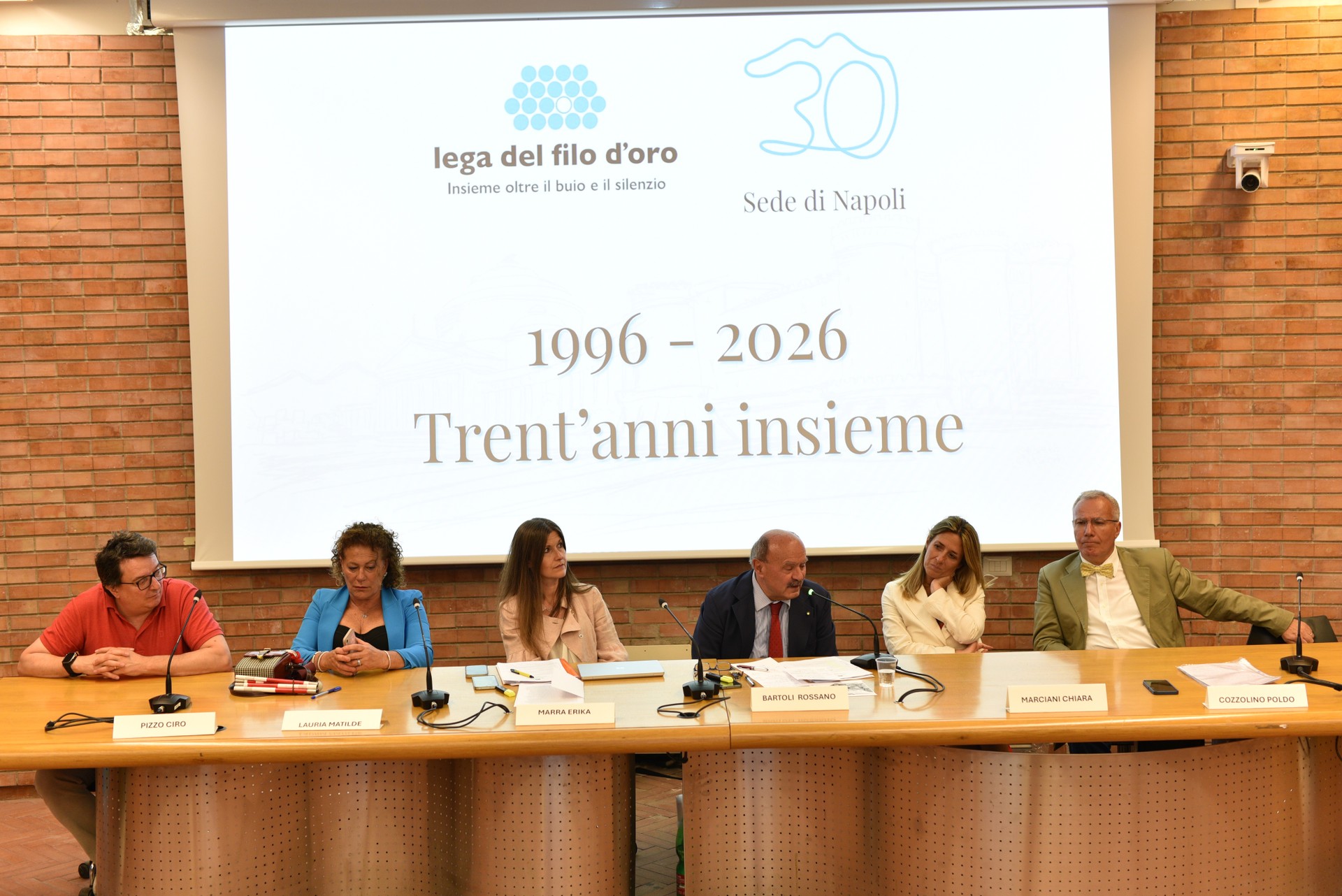Sei relatori seduti dietro un lungo tavolo con microfoni durante la celebrazione per i 30 anni della Sede Territoriale di Napoli della Lega del Filo d'Oro. Sullo sfondo, uno schermo proietta il logo “Lega del Filo d’Oro – sede di Napoli” con la scritta “1996–2026 Trent’anni insieme”.