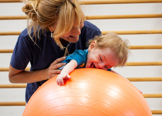 Gabriele, un bambino seguito dalla Lega del Filo d'Oro fa attività di fisioterapia con la sua fisioterapista, che lo sorregge in equilibrio sdraiato su una grande palla arancione. Entrambi ridono divertiti. 