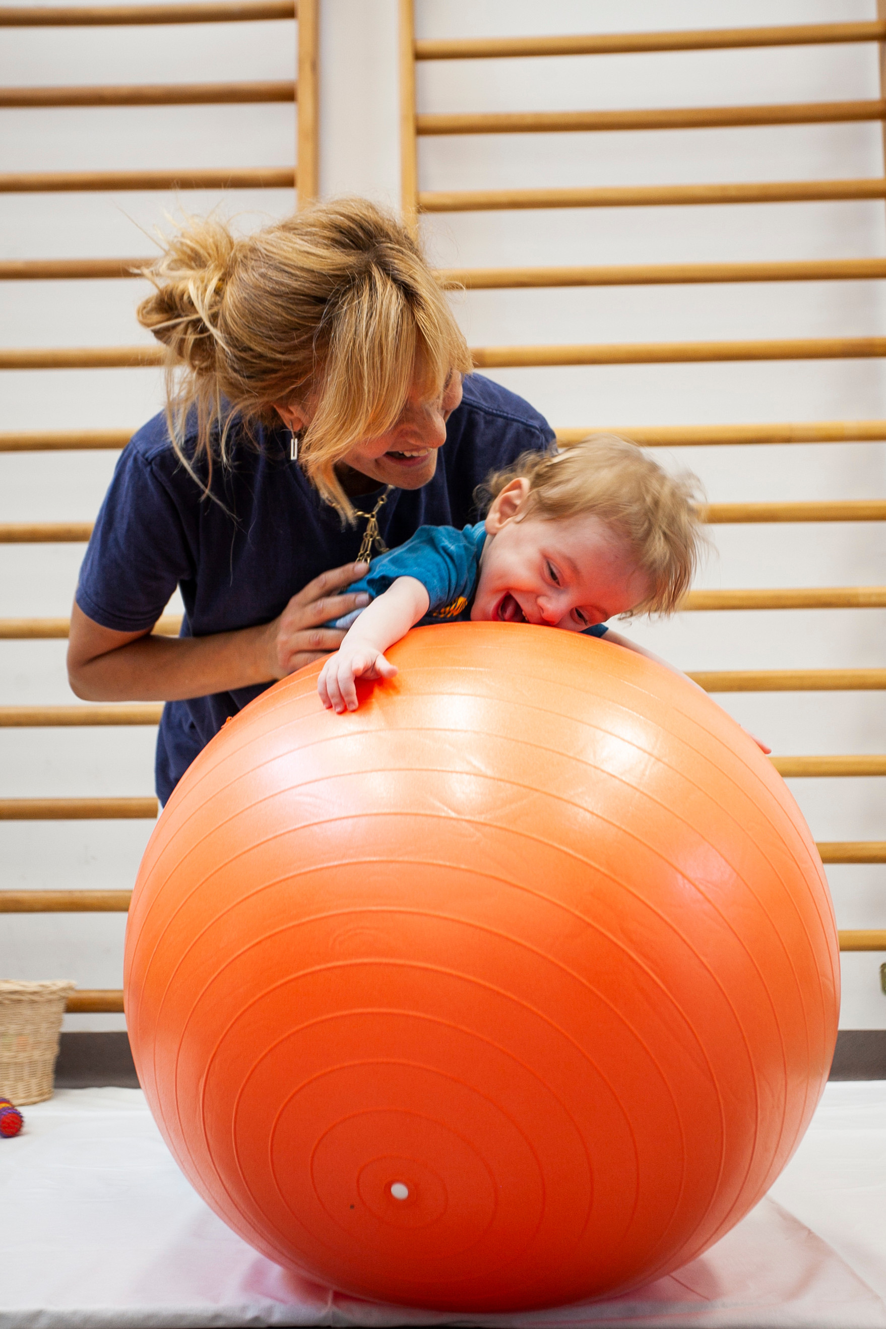 Gabriele, un bambino seguito dalla Lega del Filo d'Oro fa attività di fisioterapia con la sua fisioterapista, che lo sorregge in equilibrio sdraiato su una grande palla arancione. Entrambi ridono divertiti. 