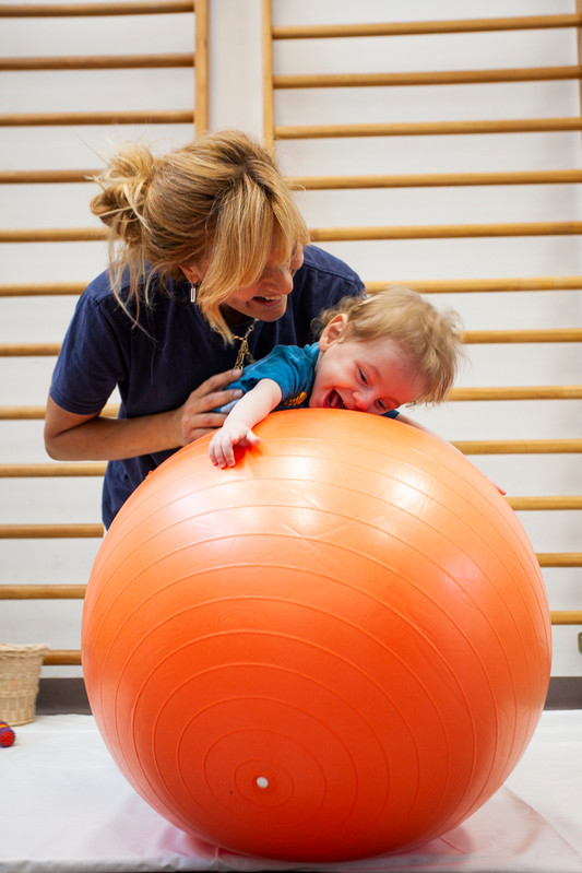 Gabriele, un bambino seguito dalla Lega del Filo d'Oro fa attività di fisioterapia con la sua fisioterapista, che lo sorregge in equilibrio sdraiato su una grande palla arancione. Entrambi ridono divertiti. 