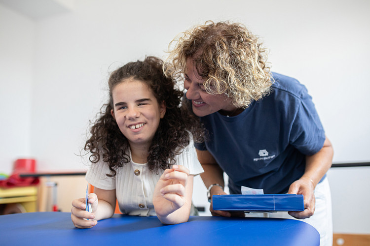 Giulia, bambina utente della Lega del Filo d’Oro, è appoggiata a un tavolo mentre tiene in mano un biglietto e scherza con un’operatrice.
