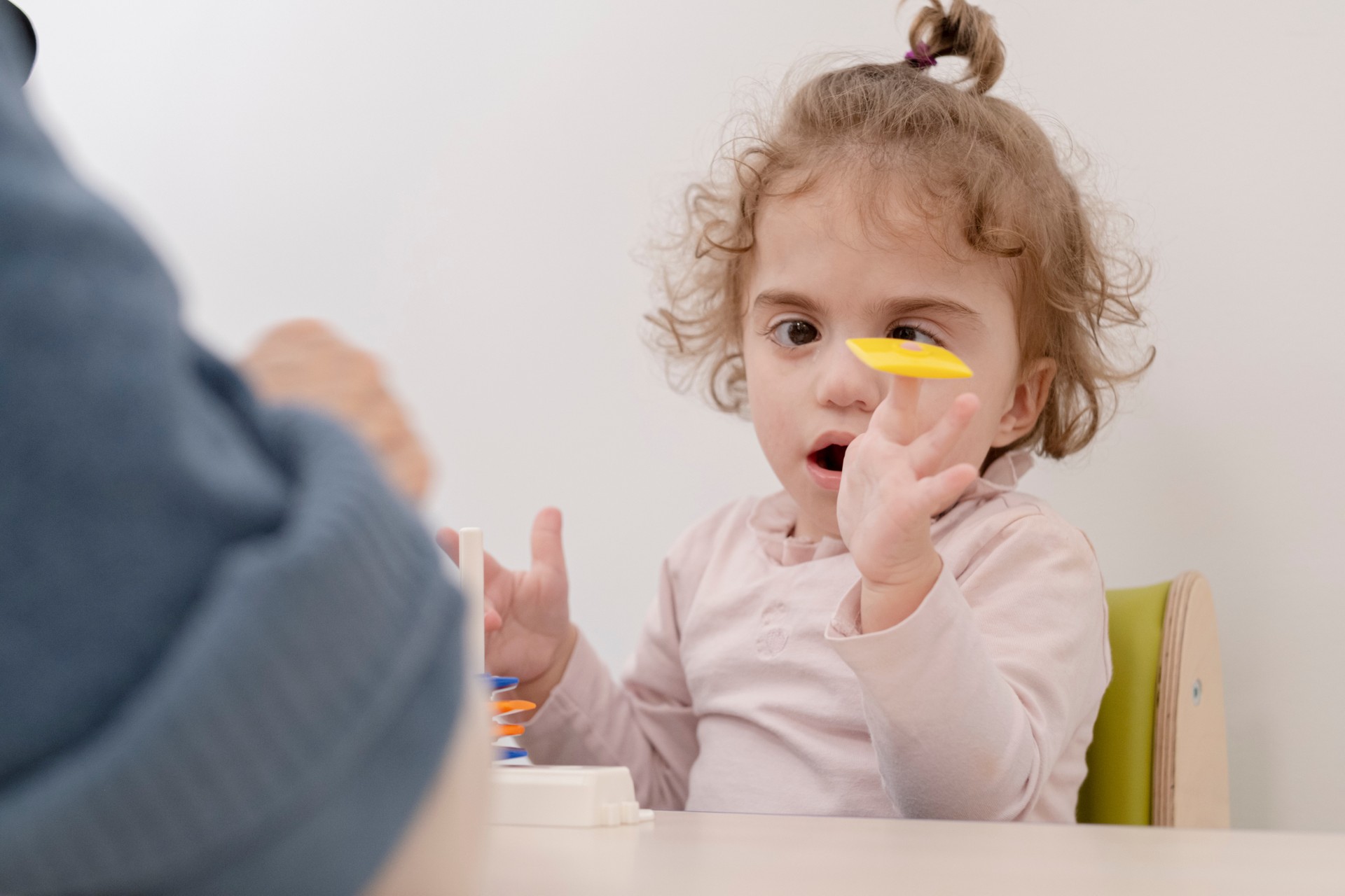 Benedetta è seduta e sta giocando con la sua educatrice. Sul tavolo davanti a lei ci sono varie forme, e lei si diverte a tenerne una in equilibro sul dito della mano sinistra