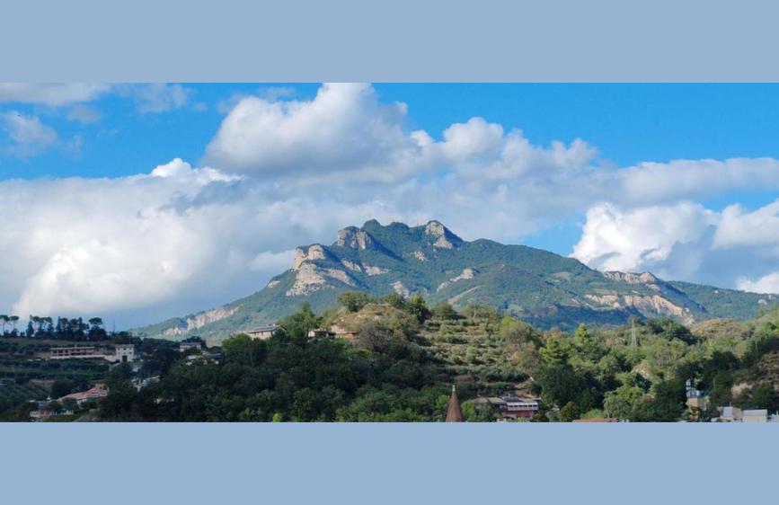 Panorama montano con cielo azzurro e nuvole, colline ricoperte di alberi e abitazioni sparse in primo piano. 