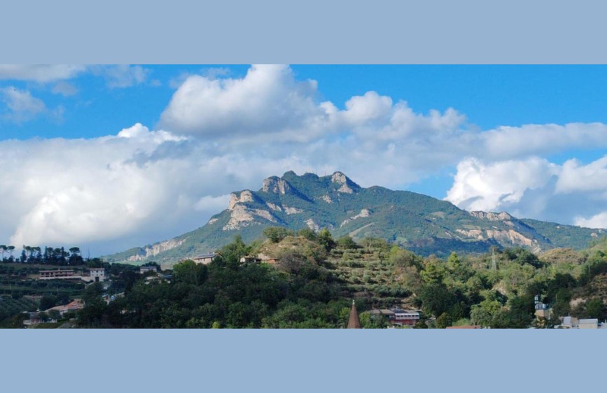 Panorama montano con cielo azzurro e nuvole, colline ricoperte di alberi e abitazioni sparse in primo piano. 