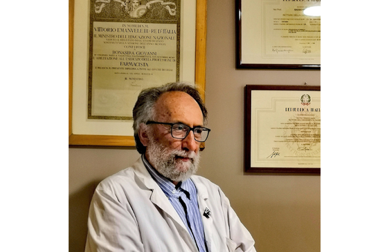 La foto ritrae un uomo con barba e occhiali, che indossa un camice bianco sopra una camicia a righe, mentre guarda leggermente verso destra. Dietro di lui, appesi a una parete, si vedono diversi diplomi o certificati incorniciati, uno dei quali sembra essere una pergamena ufficiale italiana.