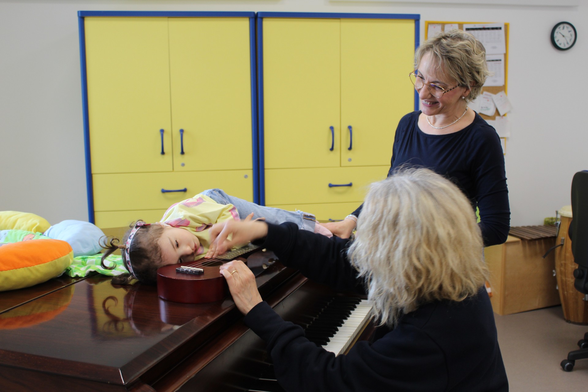 Una bambina ospite della Lega del Filo d’Oro è distesa su un pianoforte a coda durante un’attività di musicoterapia. Accanto a lei la musicoterapeuta, seduta di tre quarti e non visibile in volto, suona una chitarra appoggiata vicino alla bambina. Un’educatrice osserva la scena e sorride.