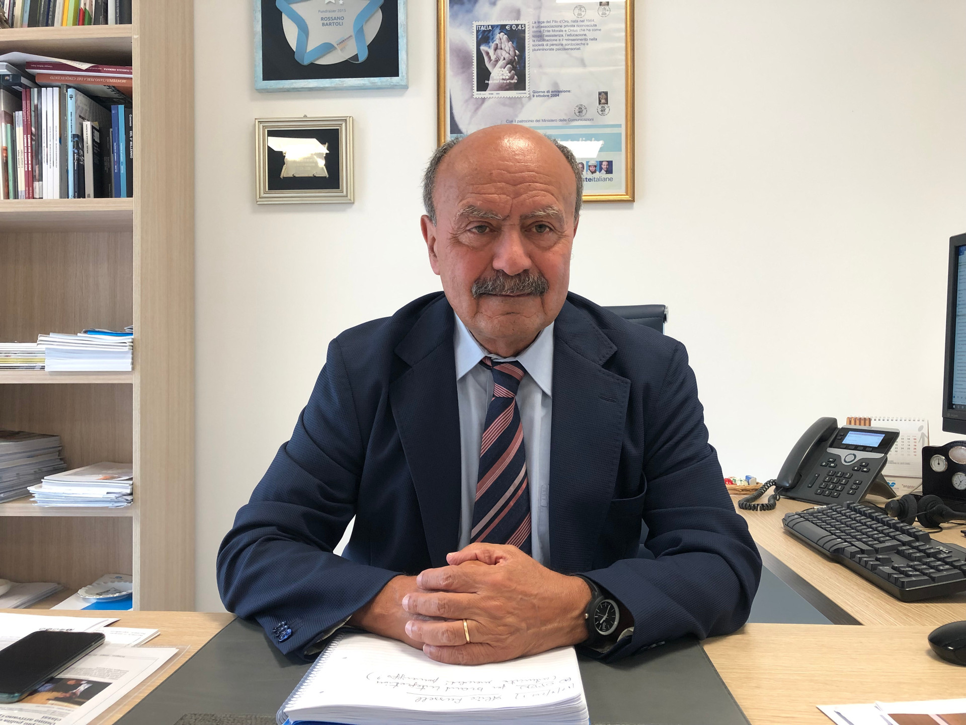 Foto a mezzo busto di Rossano Bartoli, Presidente della Lega del Filo d’Oro, seduto alla sua scrivania. Indossa giacca e cravatta.