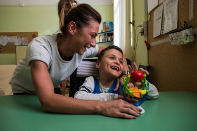 Francesco, un bambino ospite della Lega del Filo d'Oro, gioca con un giocattolo. Un'operatrice lo abbraccia e ride insieme a lui.