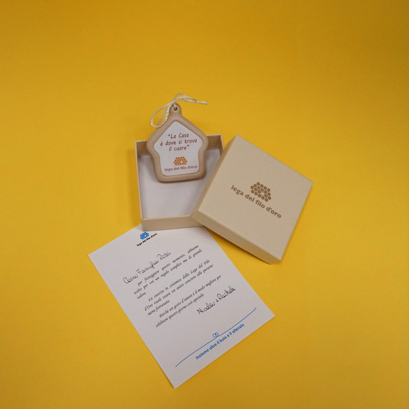 Bomboniera su sfondo giallo, composta da una casetta in ceramica appendibile, con la scritta "La casa è dove si trova il cuore", scatolina con logo della Lega del Filo d'Oro e letterina di accompagnamento personalizzata