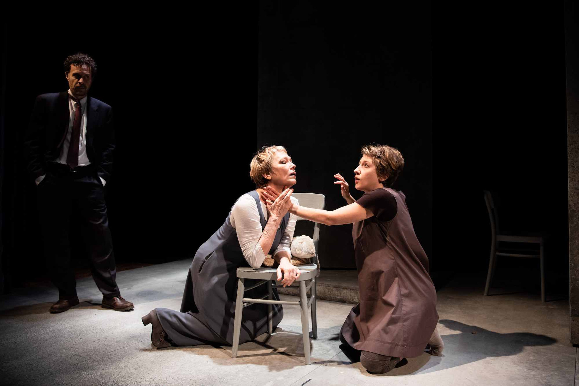  Una scena della pièce teatrale Anna dei Miracoli con due donne inginocchiate una di fronte l'altra. Sullo sfondo c'è un uomo in giacca e cravatta