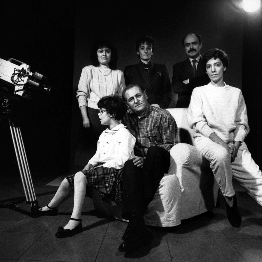 Foto in bianco e nero, scattata sul set di una campagna promozionale nel 1989. Si vede la macchina da presa e un gruppo di persone: tre sono sedute su una poltrona e sono una donna, Renzo Arbore e una bambina sordocieca; altre tre sono in piedi dietro di loro, si tratta di due donne e Rossano Bartoli.