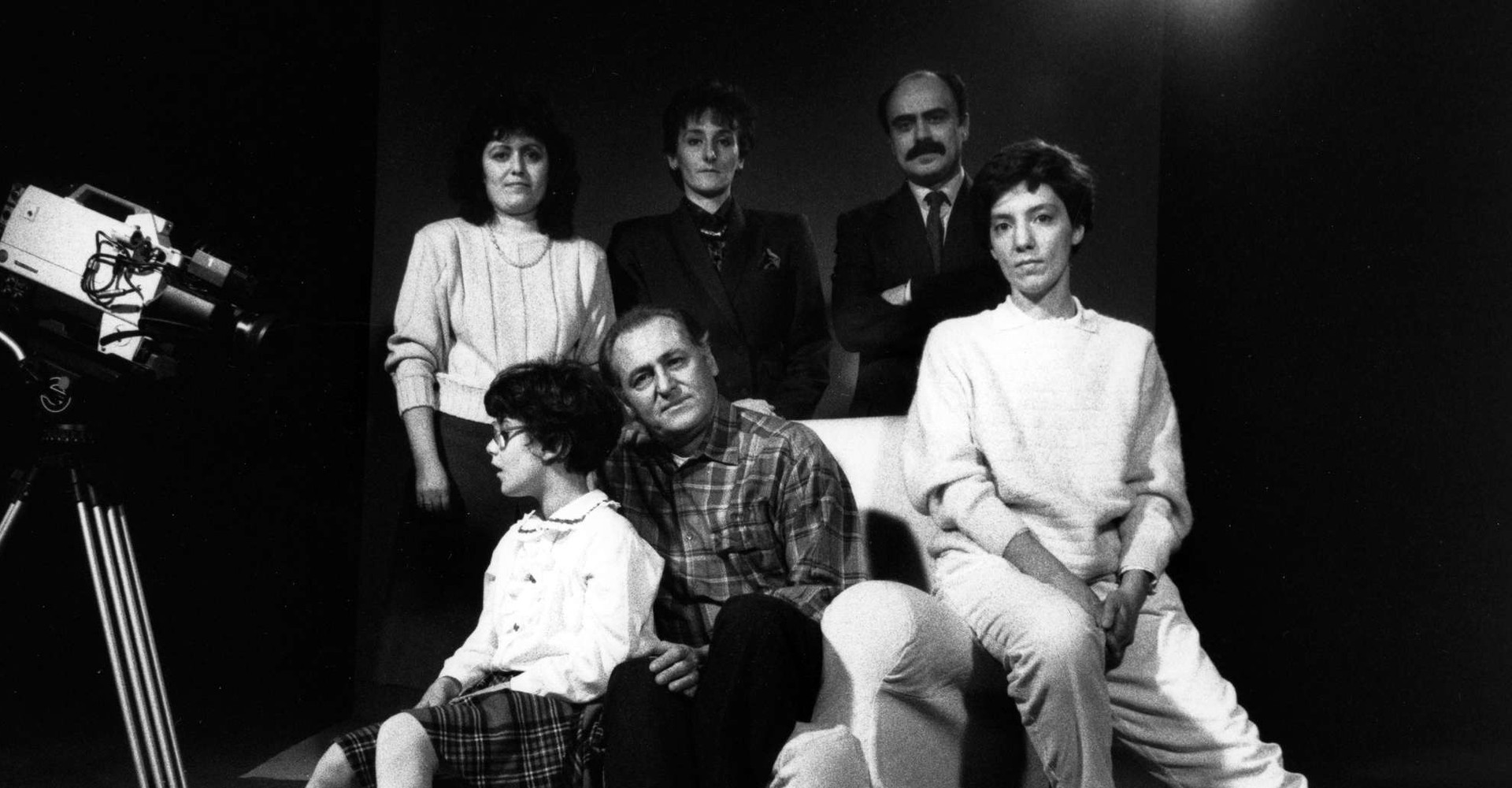 Foto in bianco e nero, scattata sul set di una campagna promozionale nel 1989. Si vede la macchina da presa e un gruppo di persone: tre sono sedute su una poltrona e sono una donna, Renzo Arbore e una bambina sordocieca; altre tre sono in piedi dietro di loro, si tratta di due donne e Rossano Bartoli.