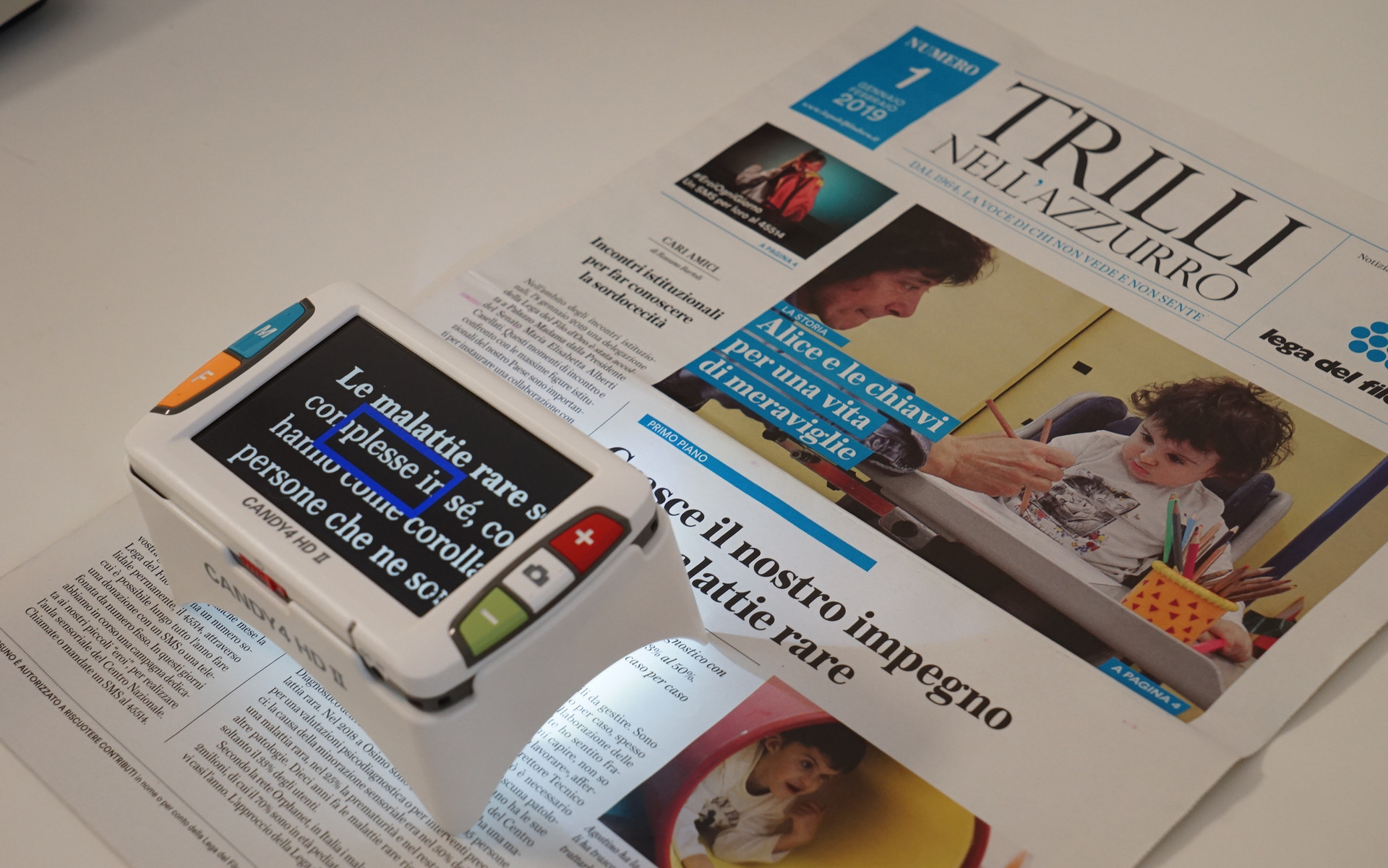 Un video ingranditore è poggiato su un numero di Trilli nell'Azzurro, il notiziario della Lega del Filo d'Oro. Sullo schermo, il testo degli articoli risulta ingrandito e il contrasto adattato per facilitare la lettura a persone con residuo visivo