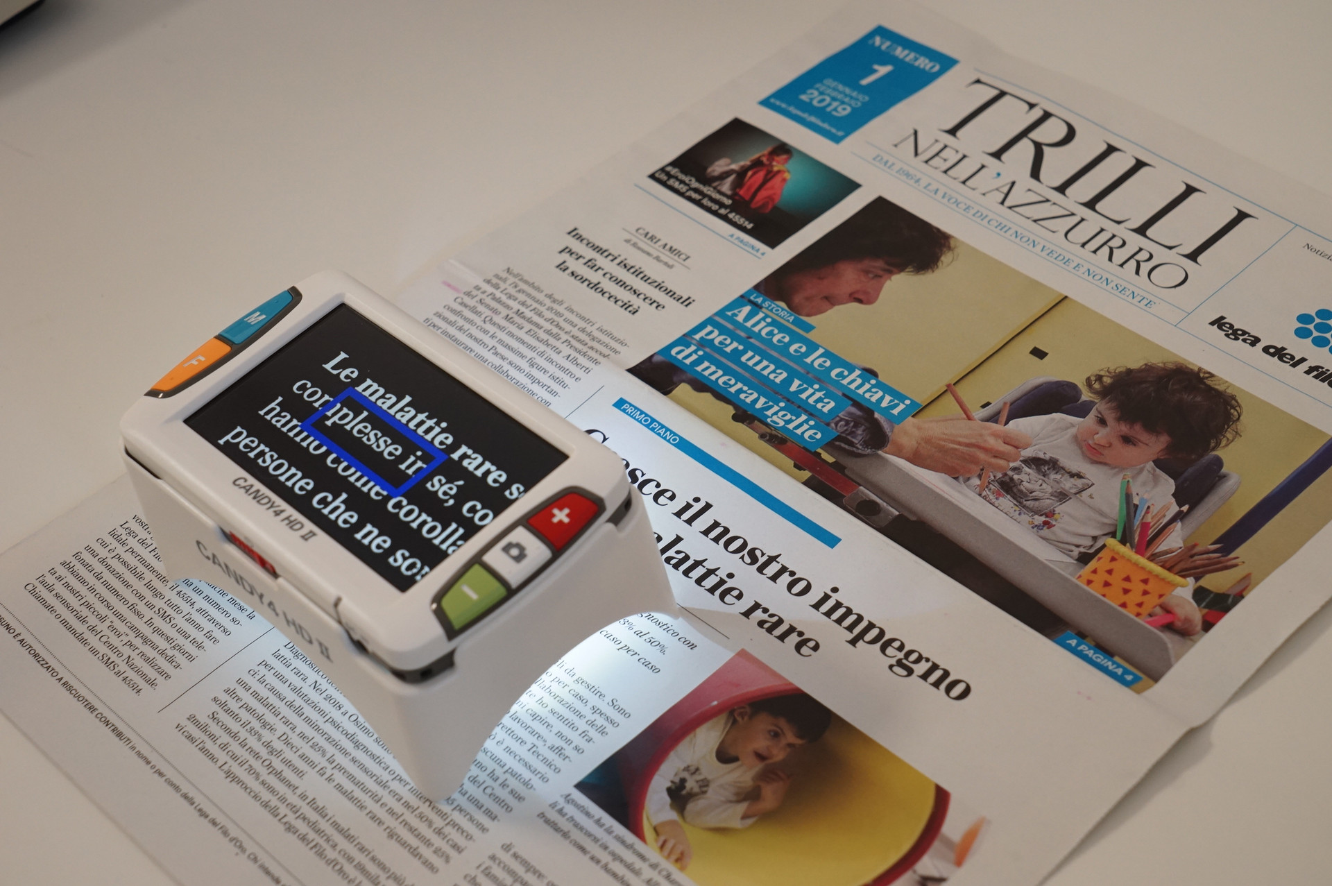 Un video ingranditore è poggiato su un numero di Trilli nell'Azzurro, il notiziario della Lega del Filo d'Oro. Sullo schermo, il testo degli articoli risulta ingrandito e il contrasto adattato per facilitare la lettura a persone con residuo visivo