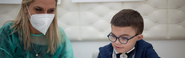 Leonardo sorride mentre l'educatrice prepara per lui un foglio con diverse immagini da associare tra loro. Al suo fianco c'è la sua educatrice, che indossa camice monouso e mascherina ffp2