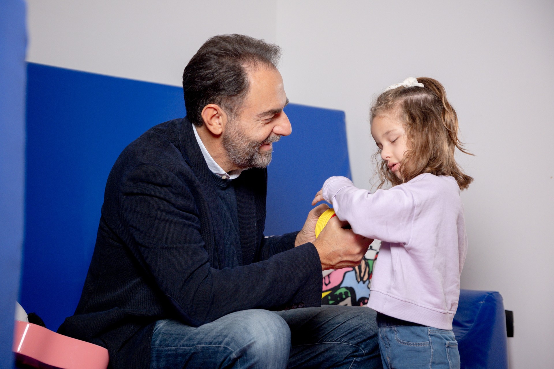 Il testimonial del 5x1000 Neri Marcorè è insieme a Chiara: lei sta giocando con la fisarmonica gialla che viene tenuta da Neri, che è seduto sul divano