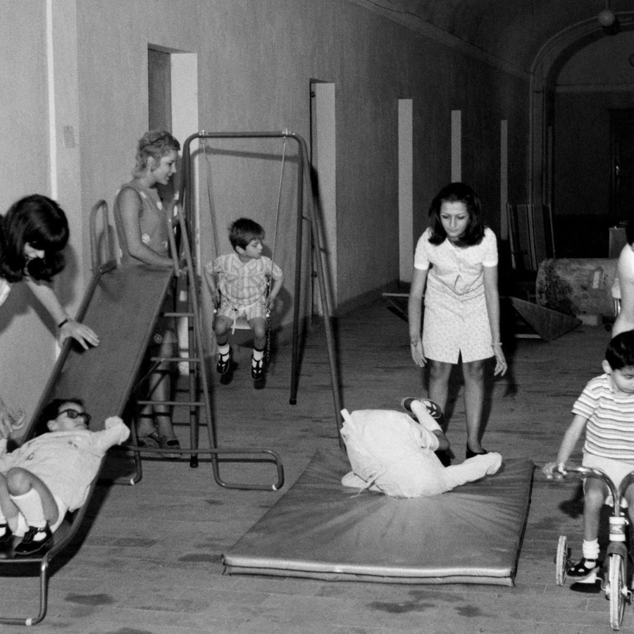Foto in bianco e nero del 1970, che ritrae giovani utenti sordociechi impegnati in attività di gioco con l'aiuto di operatrici. In un'ampia sala con tante porte, sono stati disposti uno scivolo, un'altalena, un materassino e una bicicletta. Ogni strumento è usato da un bambino, ogni bambino è seguito da un'operatrice.