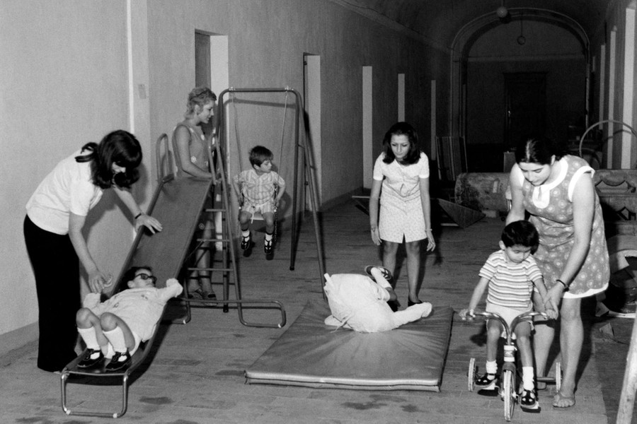 Foto in bianco e nero del 1970, che ritrae giovani utenti sordociechi impegnati in attività di gioco con l'aiuto di operatrici. In un'ampia sala con tante porte, sono stati disposti uno scivolo, un'altalena, un materassino e una bicicletta. Ogni strumento è usato da un bambino, ogni bambino è seguito da un'operatrice.