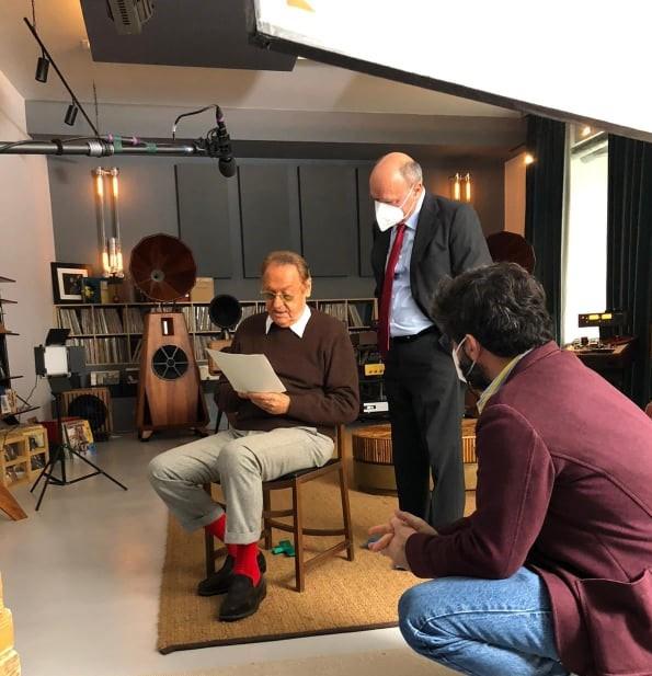 Renzo Arbore è seduto al centro della stanza e legge un foglio. Al suo fianco c'è Rosasno Bartoli. In primo piano c'è anche un altro uomo, di spalle. La stanza è abbellita con tanti mobili e soprammobili e ci sono anche le attrezzature da ripresa.