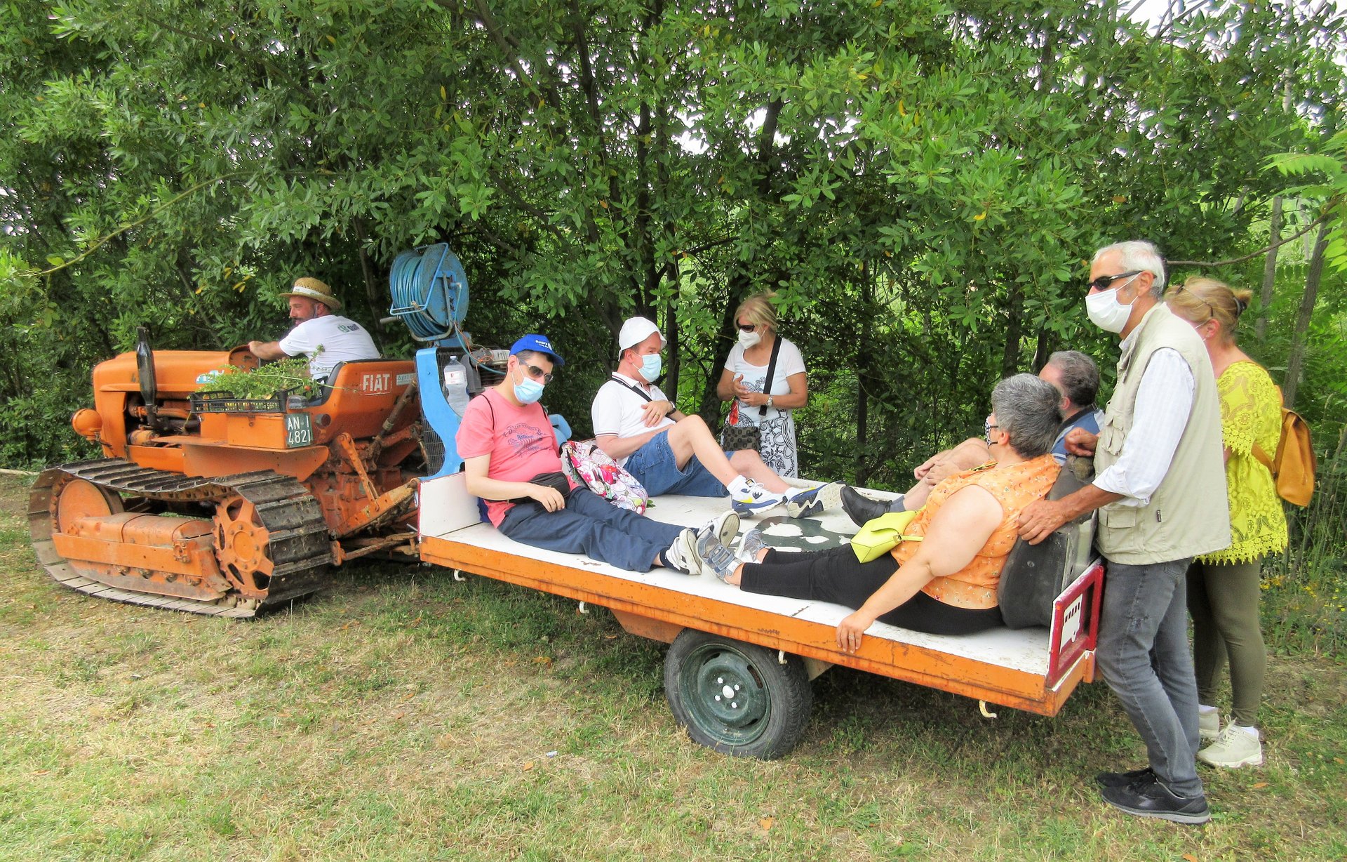 Un trattore arancione traina un carretto su cui sono seduti quattro utenti della Lega del Filo d'Oro in giro per il frutteto. Alcune persone seguono a piedi. 