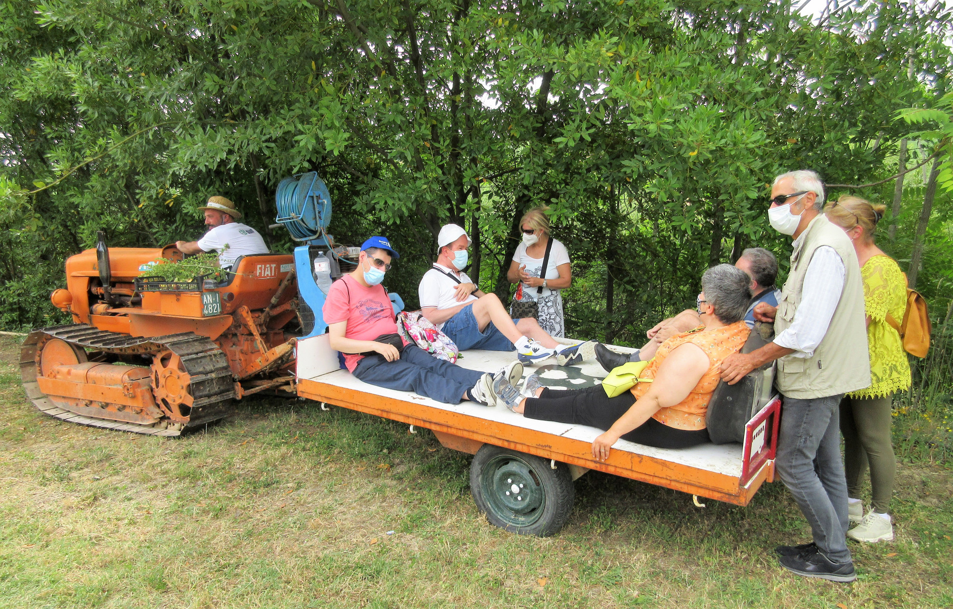 Un trattore arancione traina un carretto su cui sono seduti quattro utenti della Lega del Filo d'Oro in giro per il frutteto. Alcune persone seguono a piedi. 