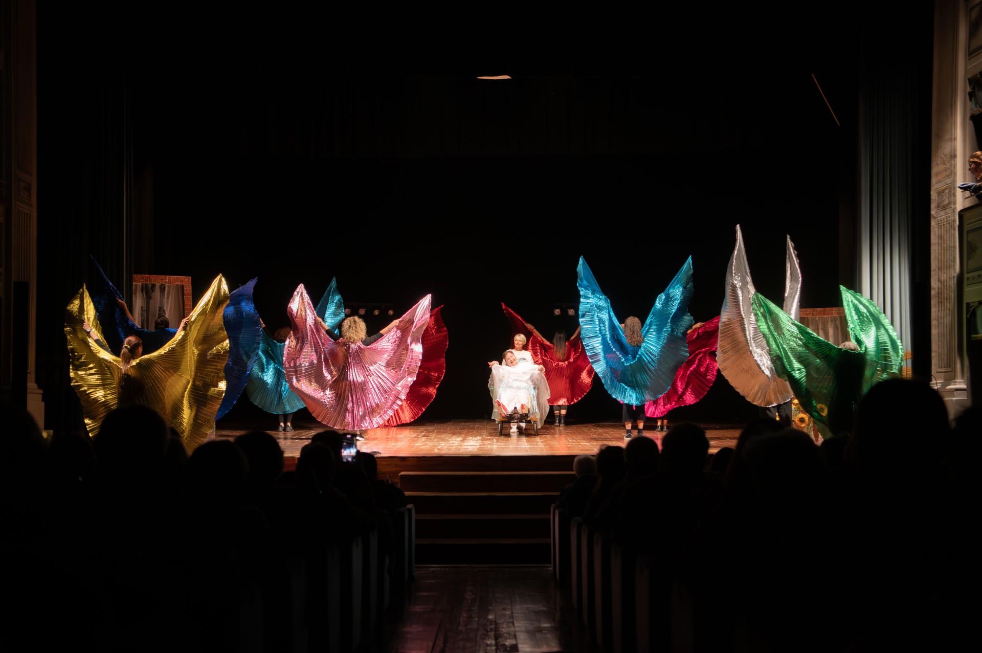 Sul palco un momento dello spettacolo “Il Condominio Sogni”, portato in scena dalla compagnia teatrale della Lega del Filo d’Oro “Il Cantiere dei Sogni”. Un gruppo di interpreti crea una coreografia con grandi ali colorate. Al centro della scena, un'ospite della Fondazione, seduta, è circondata dai movimenti armonici del cast, davanti al pubblico in sala.
