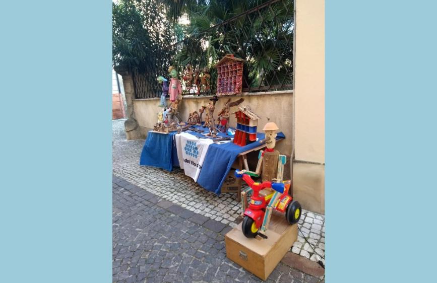 bancarella artigianale all'aperto, con un telo blu che espone la scritta "Viva del Rio". Sopra il tavolo ci sono varie sculture in legno e oggetti intagliati, mentre in primo piano si nota un grande giocattolo di legno a forma di triciclo colorato con un pupazzo seduto sopra. Lo sfondo è un vicolo pavimentato con sanpietrini e una parete chiara.