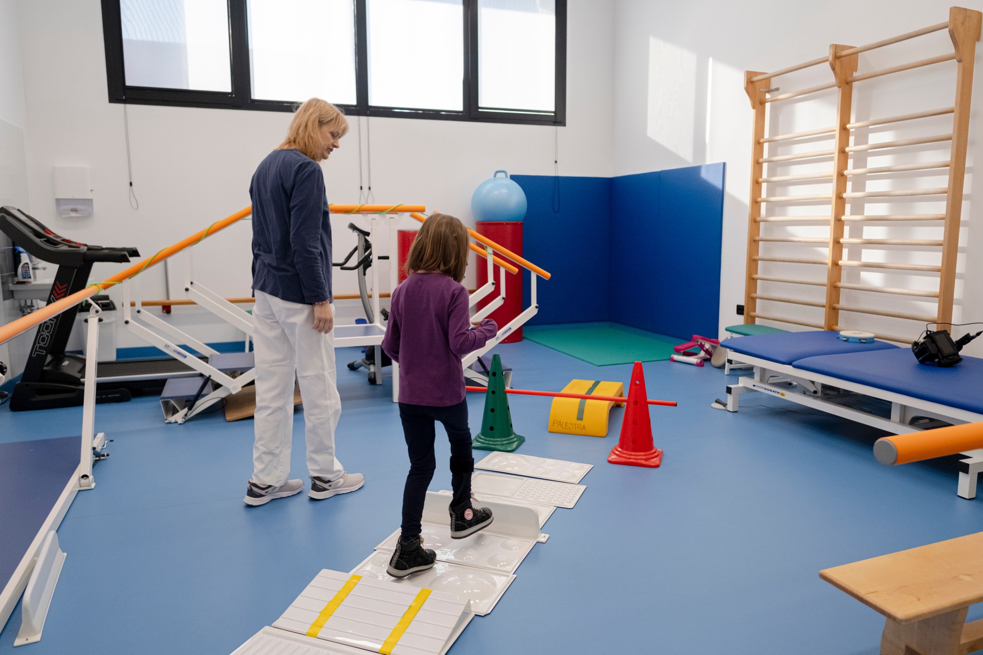 In una palestra attrezzata con parallele, lettino fisioterapico e strumenti per la riabilitazione motoria, una bambina ospite della Lega del Filo d'Oro percorre un tracciato con superfici e ostacoli predisposti per l'esercizio. Accanto a lei, una fisioterapista accompagna l'attività passo dopo passo.