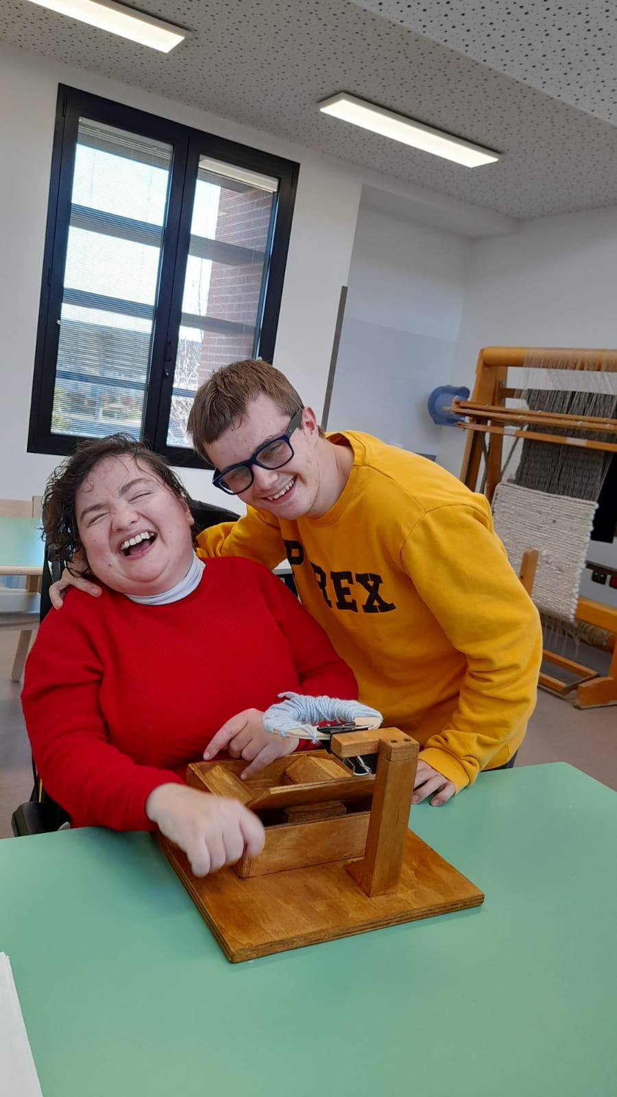 Paola e Victor, due utenti della Lega del Filo d'Oro ridono. Paola è seduta a un tavolo e lavora la maglia con un fuso per creare un pompon. Victor è in piedi e la abbraccia.