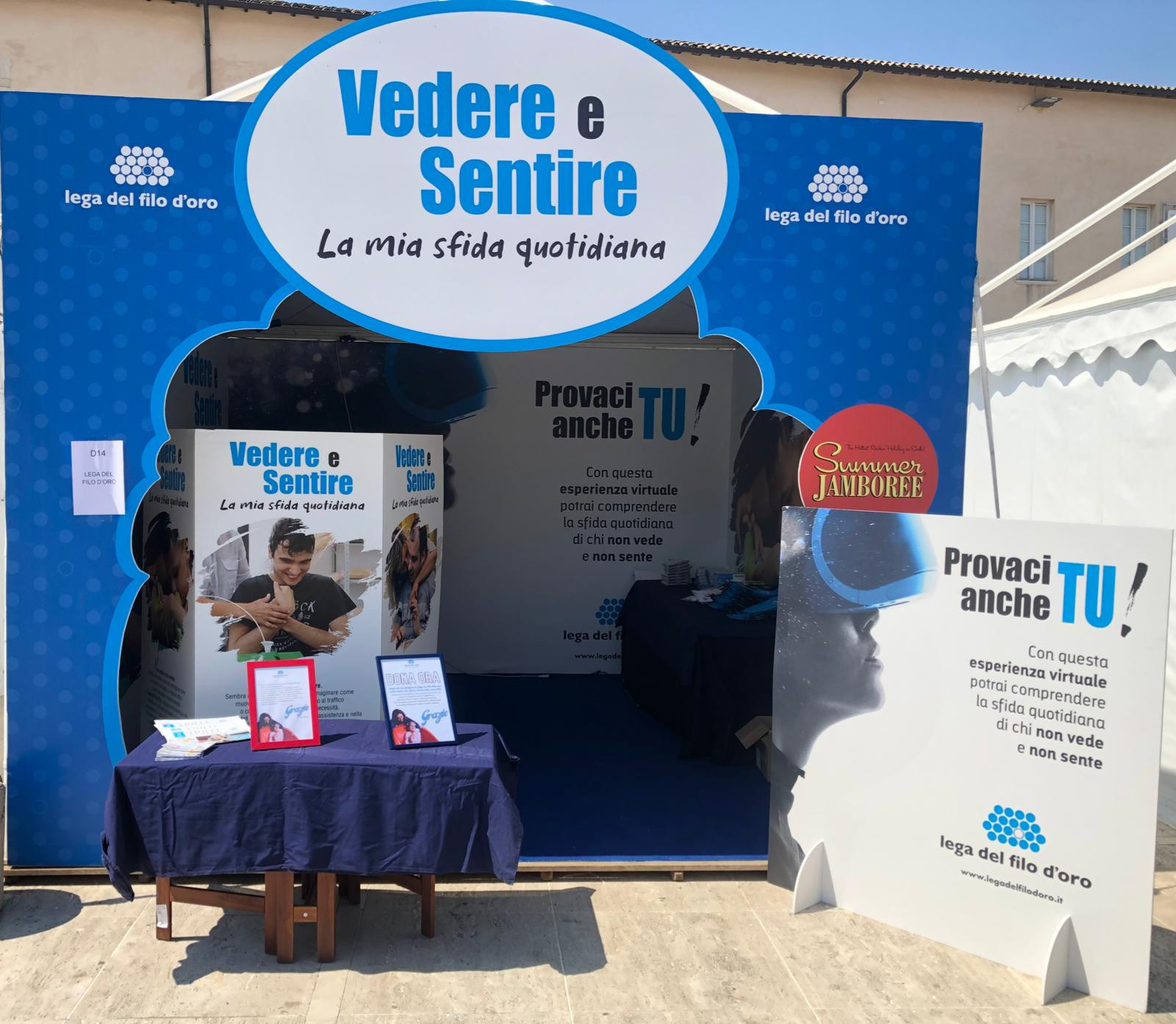 Stand azzurro della Lega del Filo d'Oro al Summer Jamboree con lo slogan “Vedere e sentire: la mia sfida quotidiana”. Un cartellone invita i visitatori a provare un’esperienza virtuale per comprendere cosa significa non vedere e non sentire.