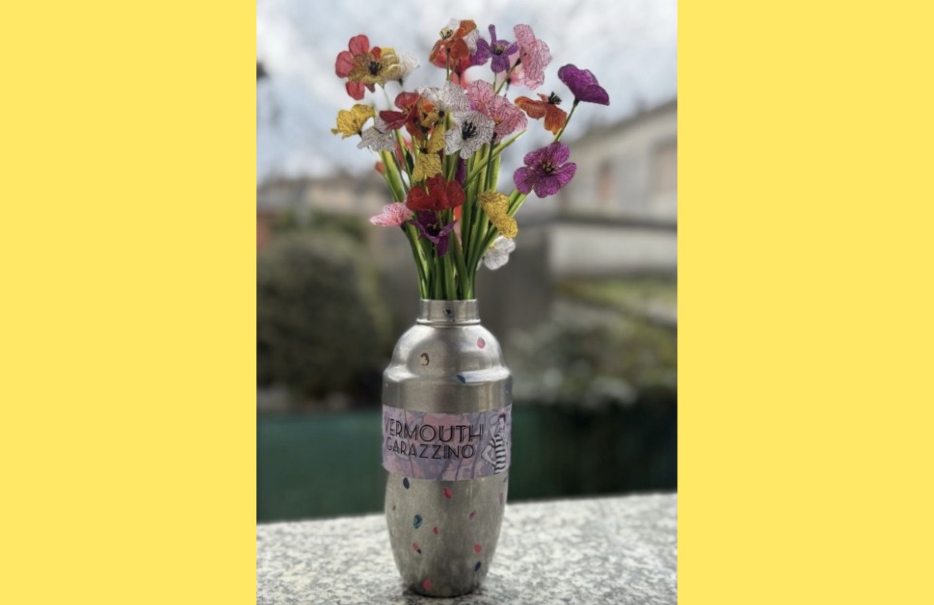 Vaso artigianale realizzato con una vecchia bottiglia di metallo decorata con pois colorati e un’etichetta con la scritta “VERMOUTH GARAZZINO”. All’interno del vaso è inserito un bouquet di fiori variopinti, probabilmente artificiali o fatti a mano, in diverse tonalità di rosso, giallo, viola e rosa. Sullo sfondo, un ambiente esterno sfocato con cespugli e edifici. La scena è incorniciata da un bordo giallo. 