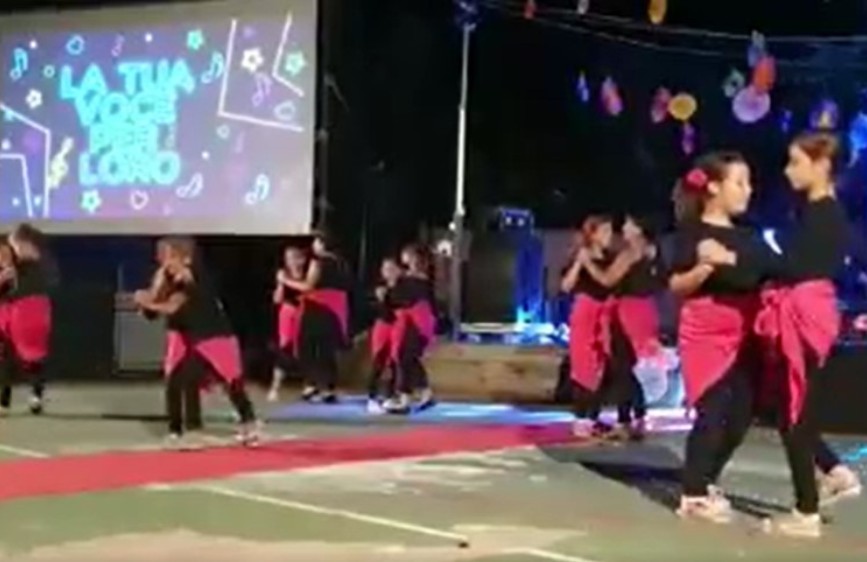L'immagine cattura una performance di danza su un palco all'aperto, probabilmente di sera. Un gruppo di bambini e bambine indossa costumi neri con dettagli fucsia, muovendosi in sincronia. Sullo sfondo, un grande schermo proietta la scritta luminosa "LA TUA VOCE PER LORO", mentre decorazioni colorate pendono sopra il palco, suggerendo un evento musicale o di beneficenza.