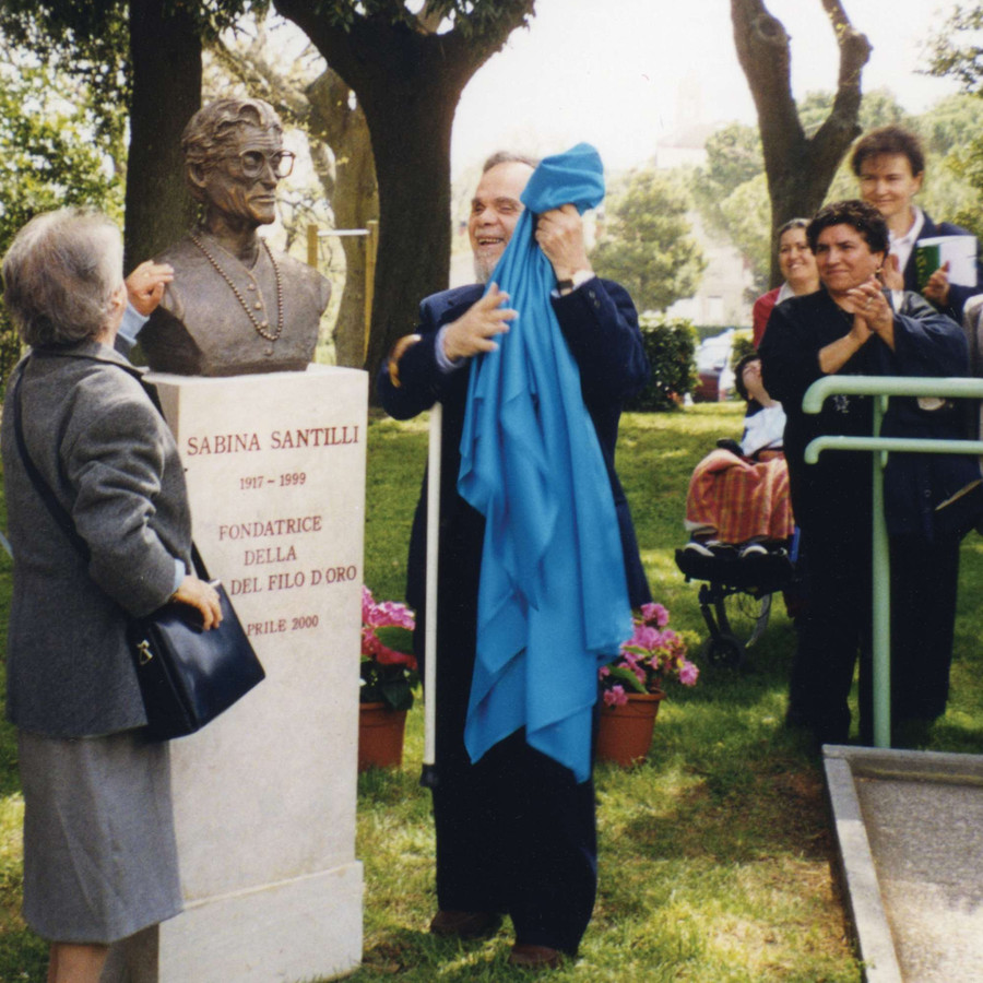 Due persone hanno appena tolto un velo azzurro dal busto dedicato a Sabina Santilli. Presente una piccola folla che applaude.