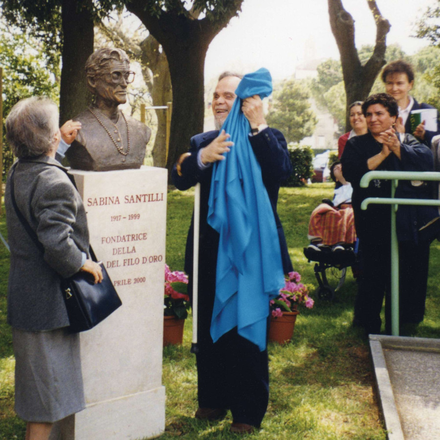 Due persone hanno appena tolto un velo azzurro dal busto dedicato a Sabina Santilli. Presente una piccola folla che applaude.