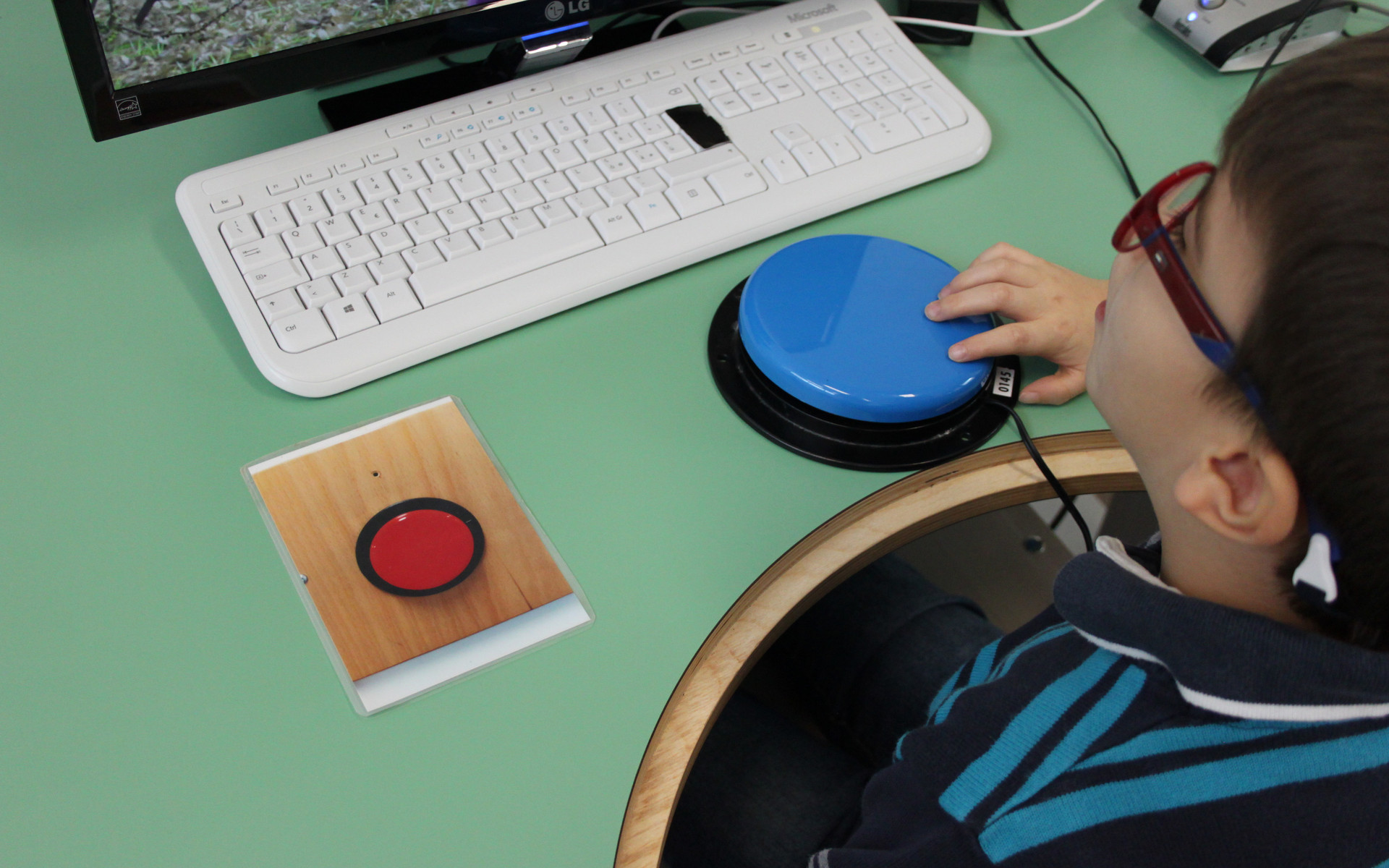 Un bambino, utente della Lega del Filo d'Oro, guarda uno schermo e preme un pulsante blu appoggiato davanti a lui per dare comandi al computer.