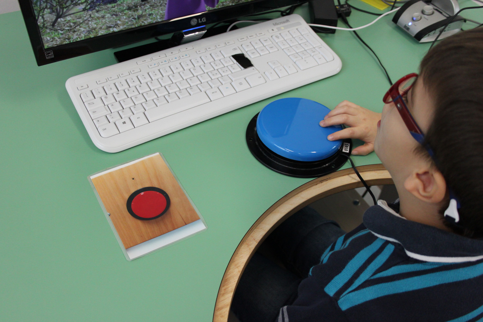 Un bambino, utente della Lega del Filo d'Oro, guarda uno schermo e preme un pulsante blu appoggiato davanti a lui per dare comandi al computer.