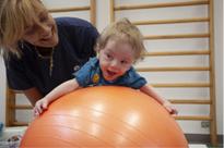 Un Bambino, utente della Lega del filo d'Oro, che sorride giocando con una palla medica di colore arancione, aiutato da un'educatrice