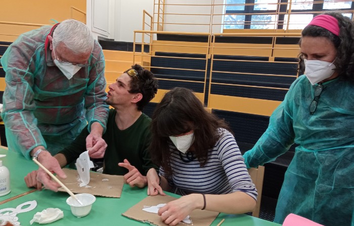 Due ospiti adulti attacano delle maschere di carta su un cartone, con il supporto dei loro educatori che indossano camice monouso e mascherina ffp2. 