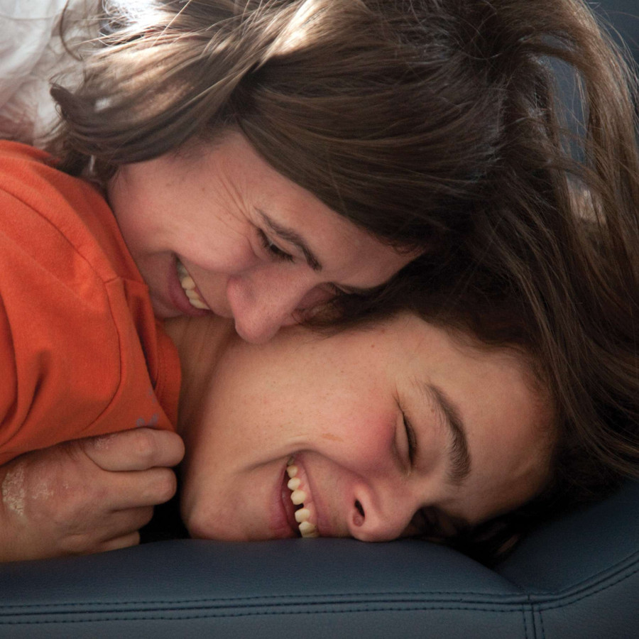Primo piano dei volti di un bambino ospite della Lega del Filo d'Oro e di una donna. Il ragazzo è appoggiato con la testa su un materasso e la donna è appoggiata su di lui e lo abbraccia. Entrambi sorridono.