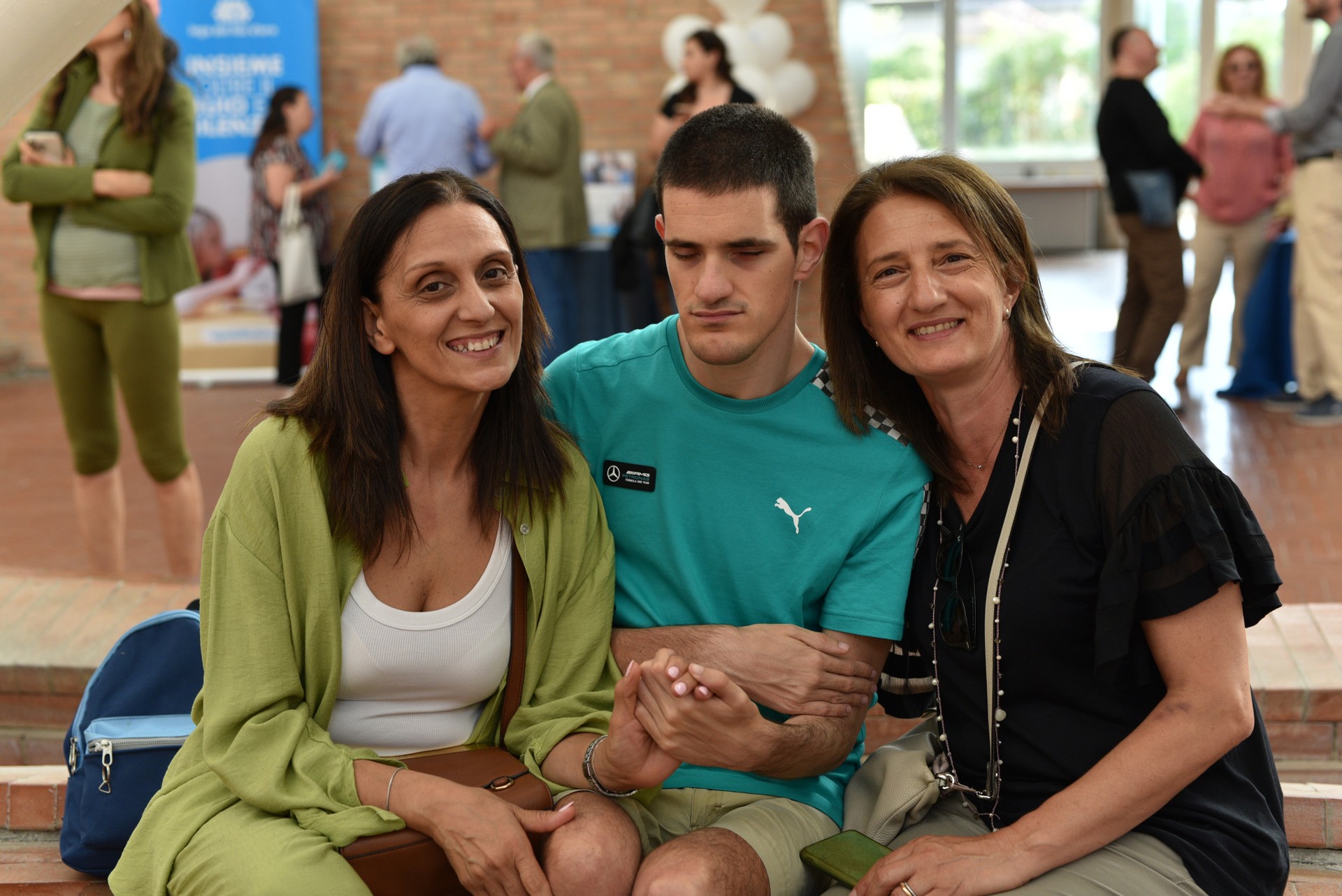 Un utente partecipa alla presentazione dei 30 anni della Sede di Napoli. E' seduto con le braccia incrociate in mezzo a due signore che lo abbraciano da dietro e sorridono in favore della telecamera