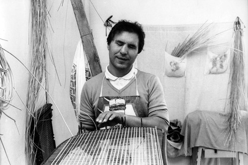 Immagine in bianco e nero degli anni '70. Un uomo sordocieco è fotograto mentre lavora ad un manufatto di vimini.