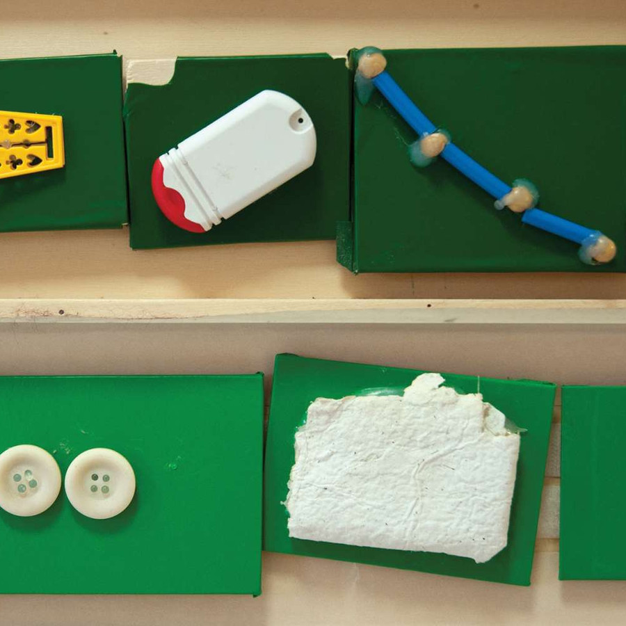 Alcuni tasselli di legno verdi e bianchi sono attaccati a un muro. Su ogni tassello è incollato un oggetto di piccole dimensioni come dei bottoni, oppure dei pezzetti di stoffa o altri materiali, delle cifre come il numero 10 o delle lettere che compongono il nome ALEX.