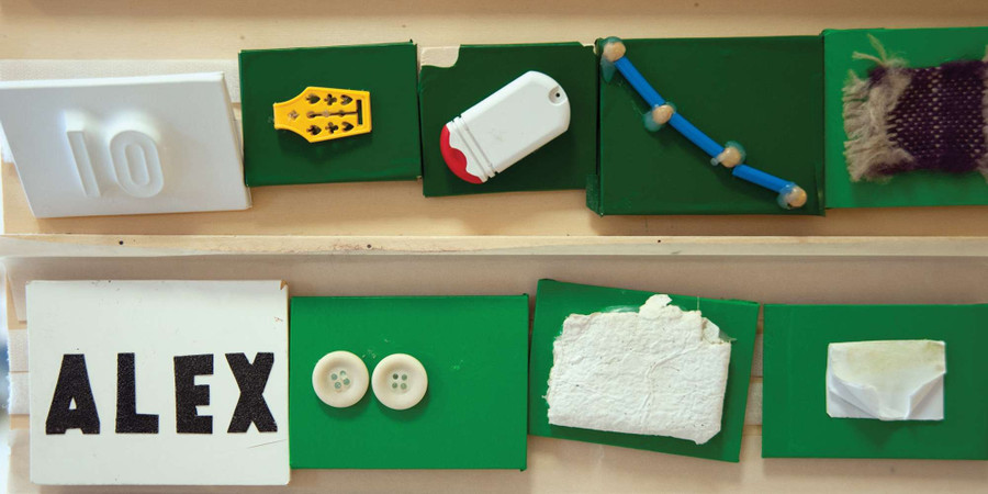Alcuni tasselli di legno verdi e bianchi sono attaccati a un muro. Su ogni tassello è incollato un oggetto di piccole dimensioni come dei bottoni, oppure dei pezzetti di stoffa o altri materiali, delle cifre come il numero 10 o delle lettere che compongono il nome ALEX.