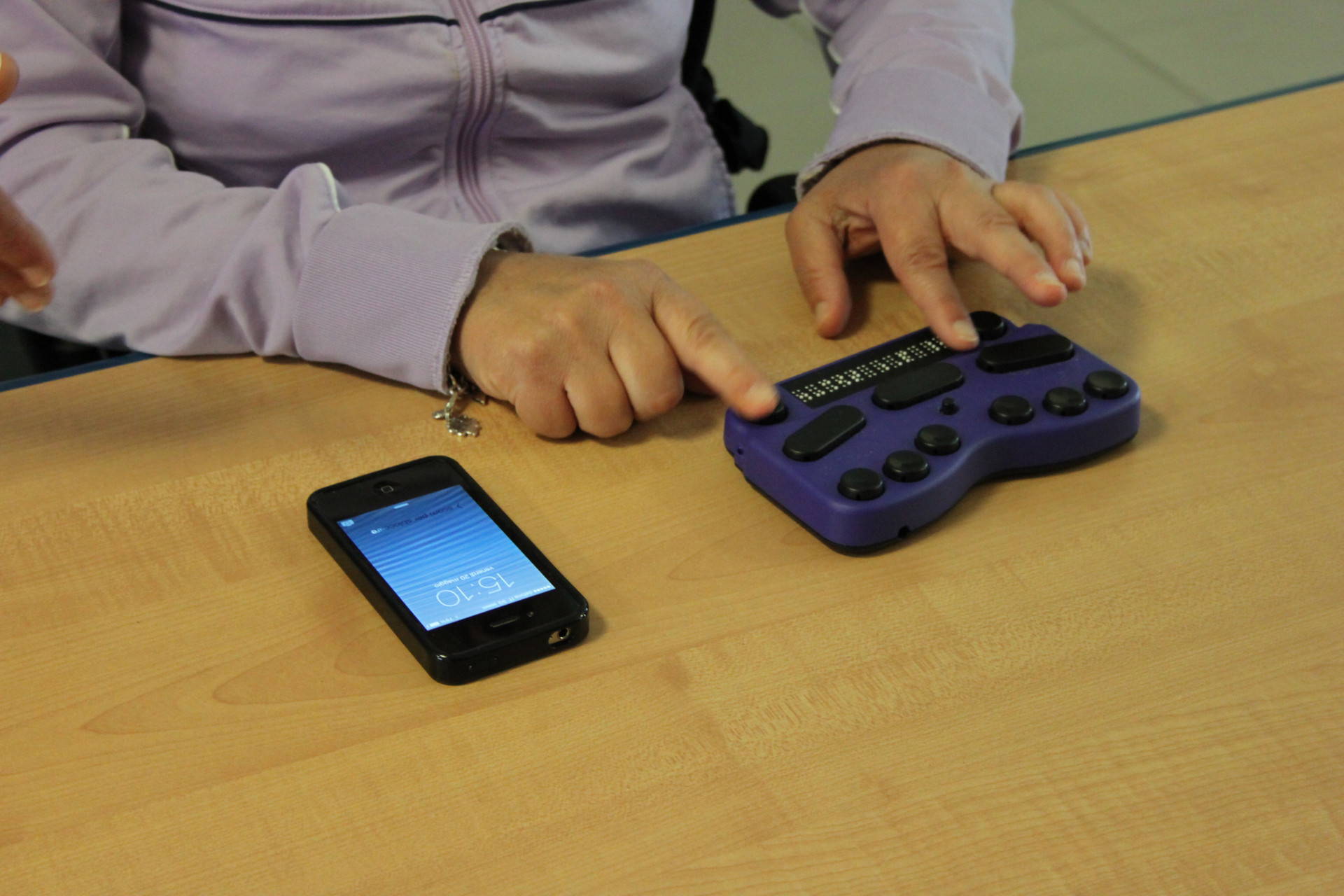 Due manni si appoggiano a una dispositivo che utilizza il braille. A fianco, sulla stessa scrivania, c'è un Iphone.