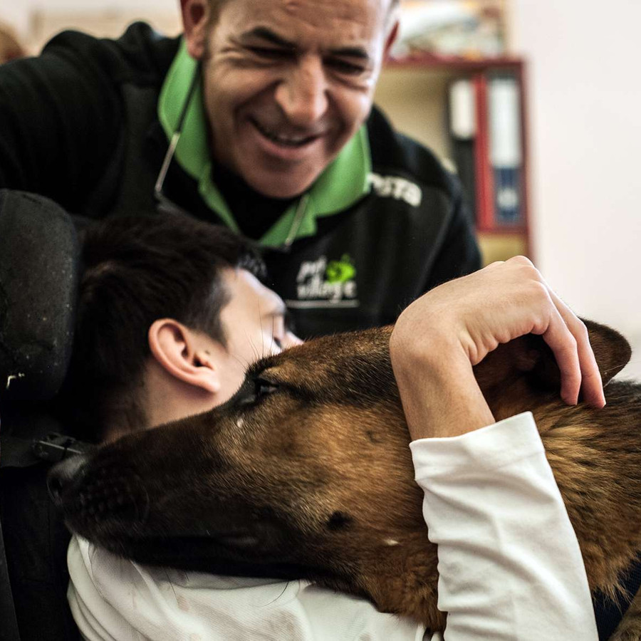 Sotto gli occhi di un operatore sorridente, un ragazzo sordocieco abbraccia un cane pastore tedesco, che appoggia affettuoso la sua testa sulla spalla dell'utente.
