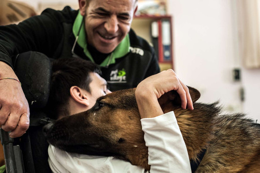 Sotto gli occhi di un operatore sorridente, un ragazzo sordocieco abbraccia un cane pastore tedesco, che appoggia affettuoso la sua testa sulla spalla dell'utente.