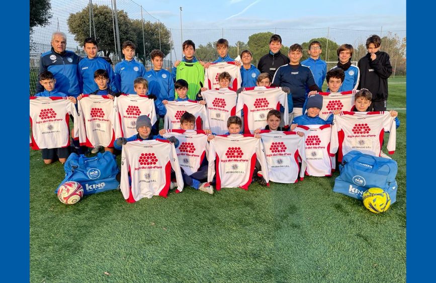 Un gruppo di giovani giocatori di calcio in maglie blu e bianche posa per una foto su un campo verde. Molti di loro tengono in mano delle maglie bianche con scritte rosse, mentre alcuni adulti sono in piedi dietro di loro.