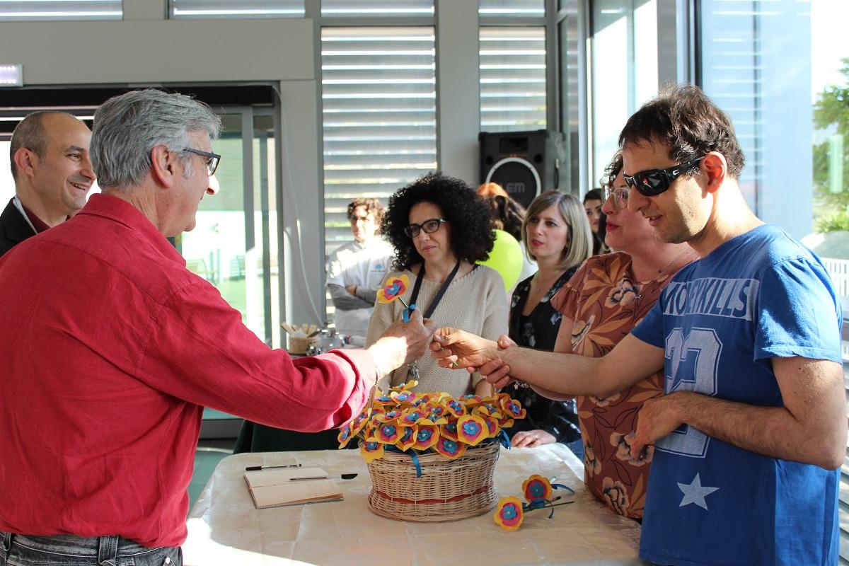 Giornata del Sostenitore - Saluti finali: un utente adulto sordocieco consegna ai sostenitori un piccolo regalo ricordo realizzato dagli utenti: una matita co in cima un fiore di carta