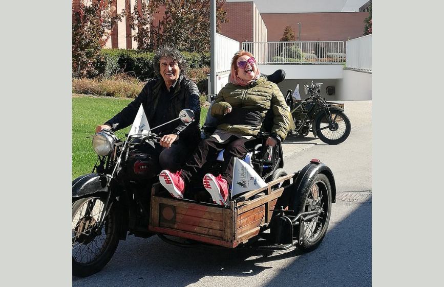Durante l’evento Sidecar 2023, l’utente Paola della Lega del Filo d’Oro è ritratta con un grande sorriso mentre si gode un giro a bordo di un sidecar d’epoca. Seduta comodamente nella carrozzina fissata al mezzo, Paola indossa un piumino verde e scarpe rosse, mentre accanto a lei l’autista guida la moto con entusiasmo. Entrambi appaiono felici, condividendo un momento di gioia e libertà all’aria aperta, all’interno di un contesto urbano soleggiato.