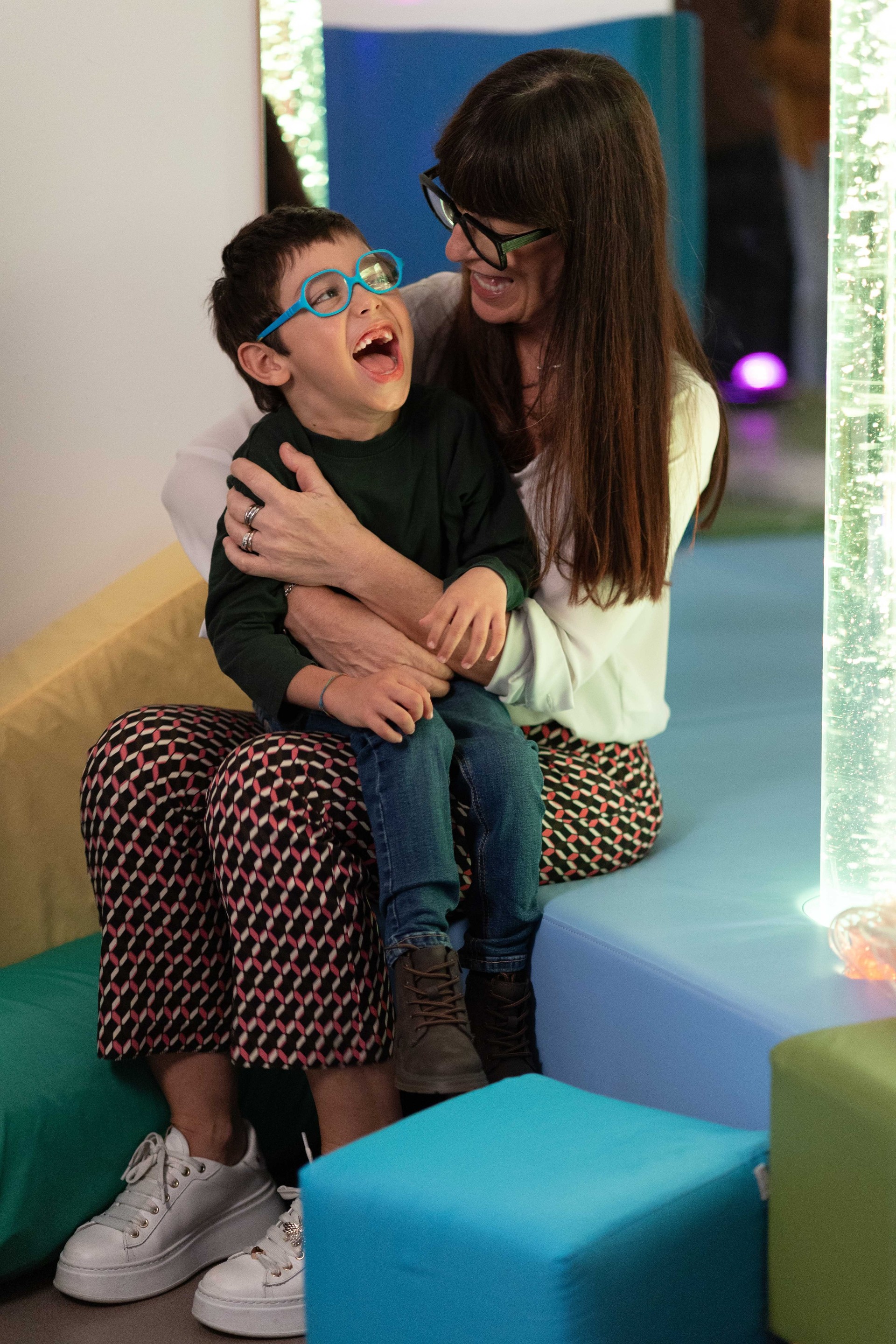 Nicola, bimbo con ipovisione e con problemi motori, seduto sulle gambe della sua educatrice, sorride nella stanza delle stimolazioni visive. Un momento di gioia in ambiente educativo.